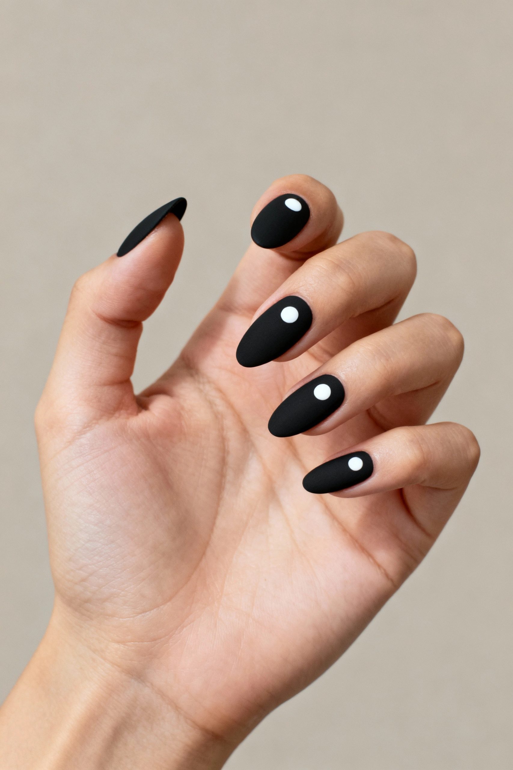 A hand with long, matte black almond-shaped nails, each decorated with a single small white dot near the cuticle.