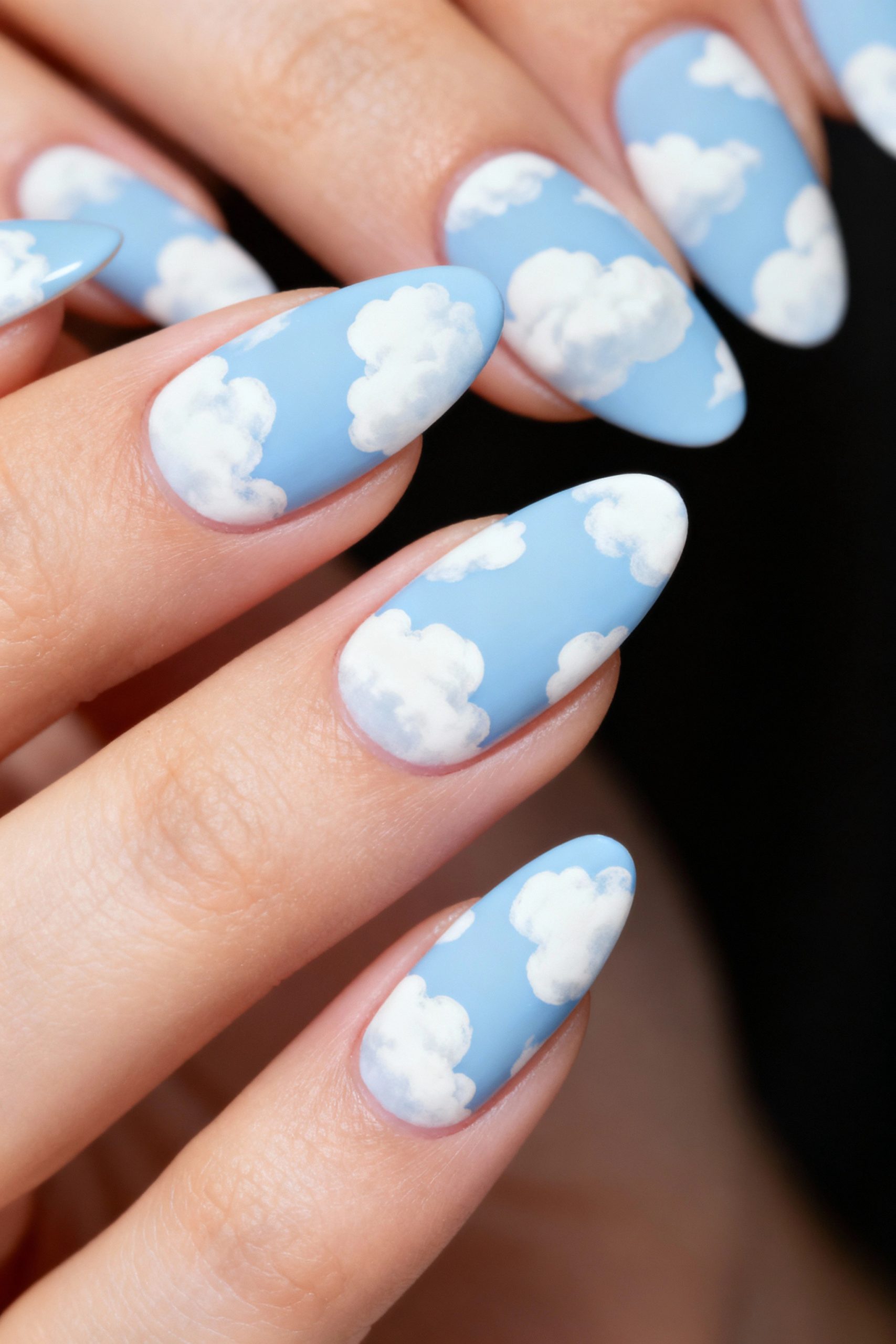 A detailed view of almond-shaped nails with a light blue base and painted white clouds, giving the effect of a daytime sky.