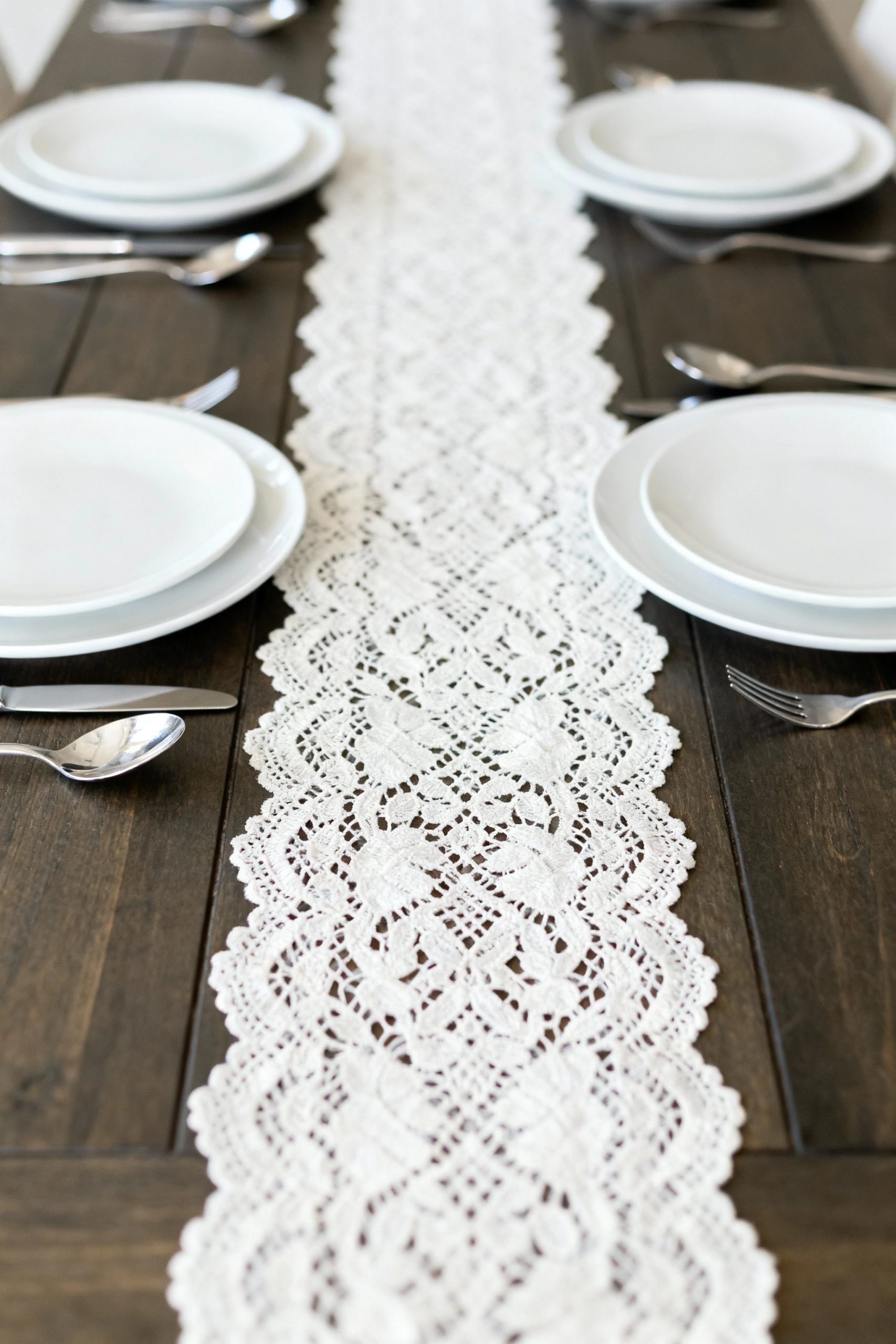 A beautifully set dining table with simple white plates and a long, elegant lace runner on a dark wood table.