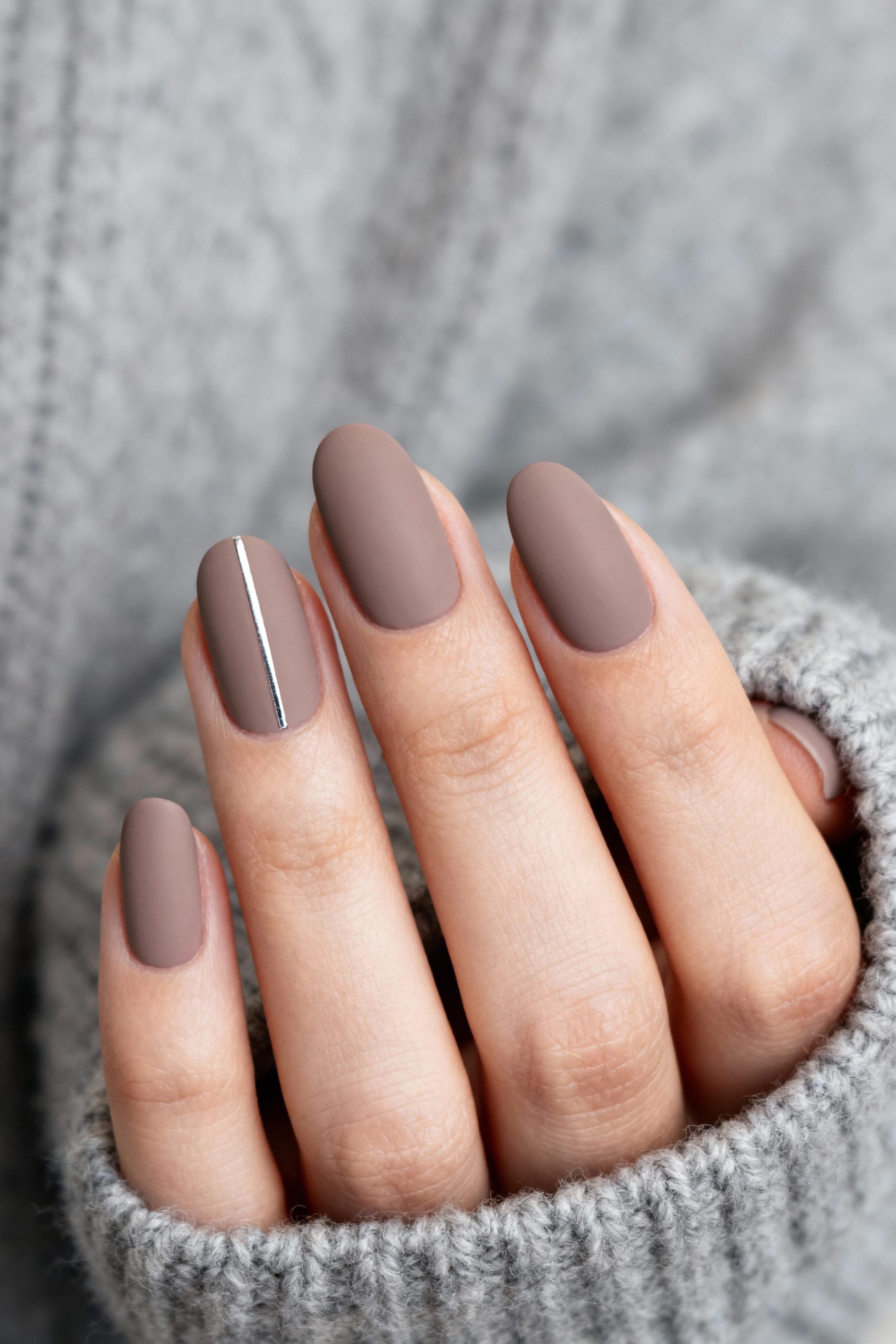 A close-up of a hand with long, oval-shaped matte taupe nails, with a single delicate silver line on one nail, resting in a cozy grey sweater sleeve.