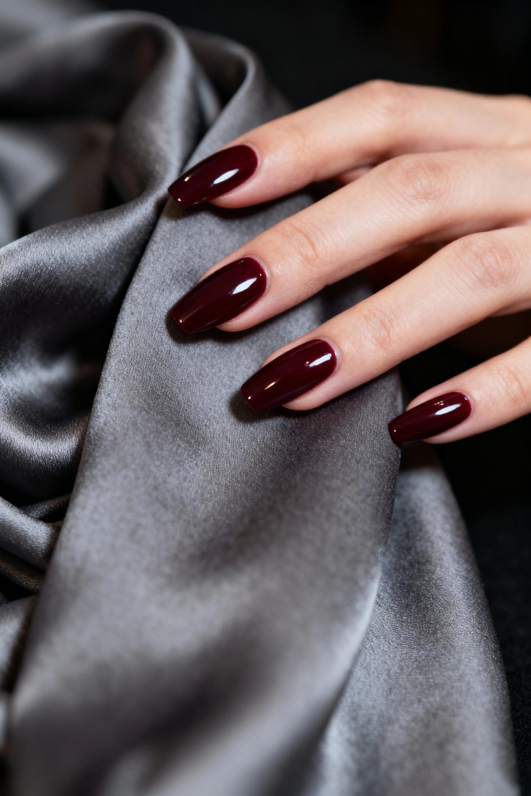 Elegant hand with long almond nails painted in a deep shiny burgundy red, resting on grey silk.