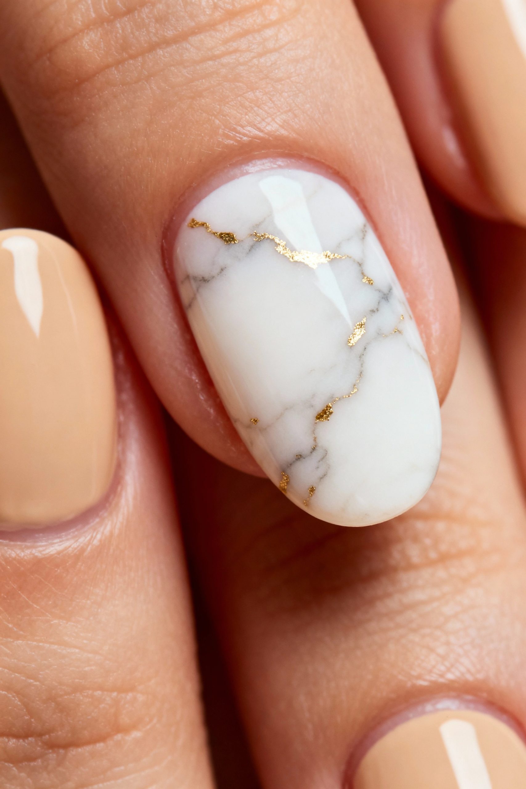 A macro photo of a stunning accent nail with a realistic white marble design and delicate gold foil veins, next to beige nails.