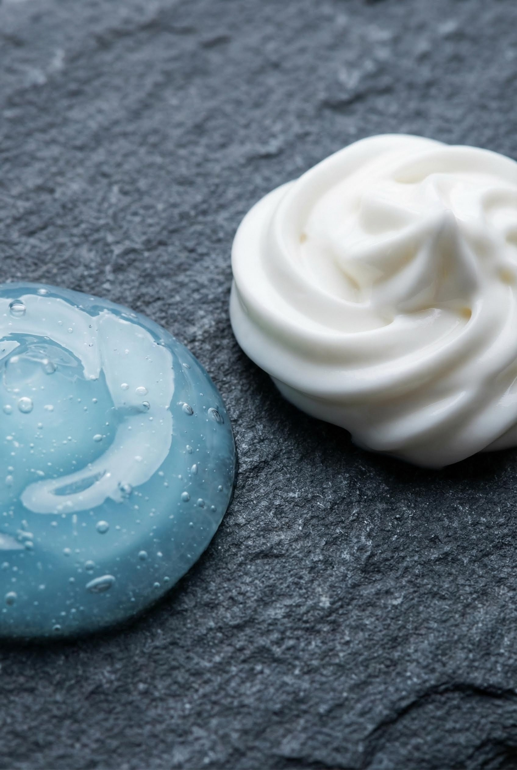 Gel Moisturizer vs. Cream Moisturizer: Best for Your Skin Type? 1 Macro photography shot of a blue transparent gel dollop next to a white rich cream swirl on a dark stone surface, showing the texture difference.