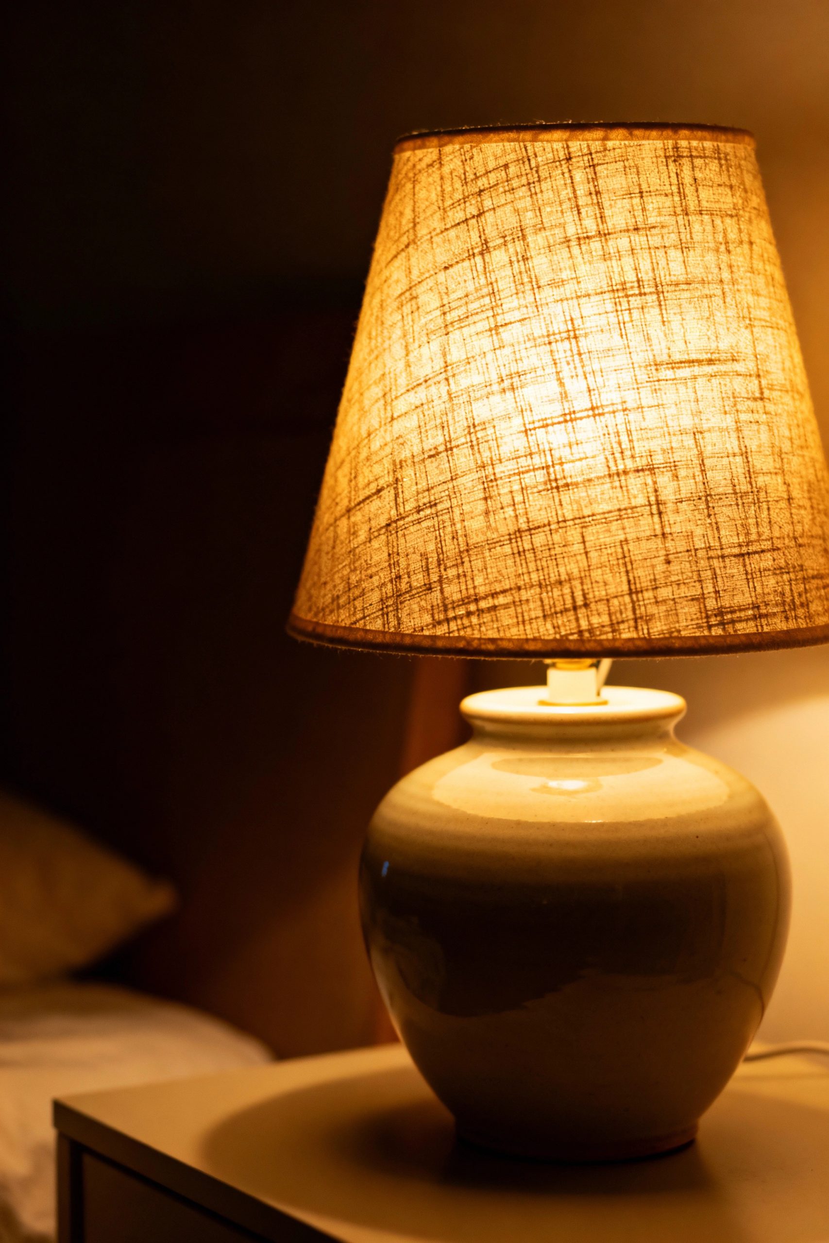 Ceramic table lamp with a textured linen shade casting a warm, cozy glow in a dimly lit room.