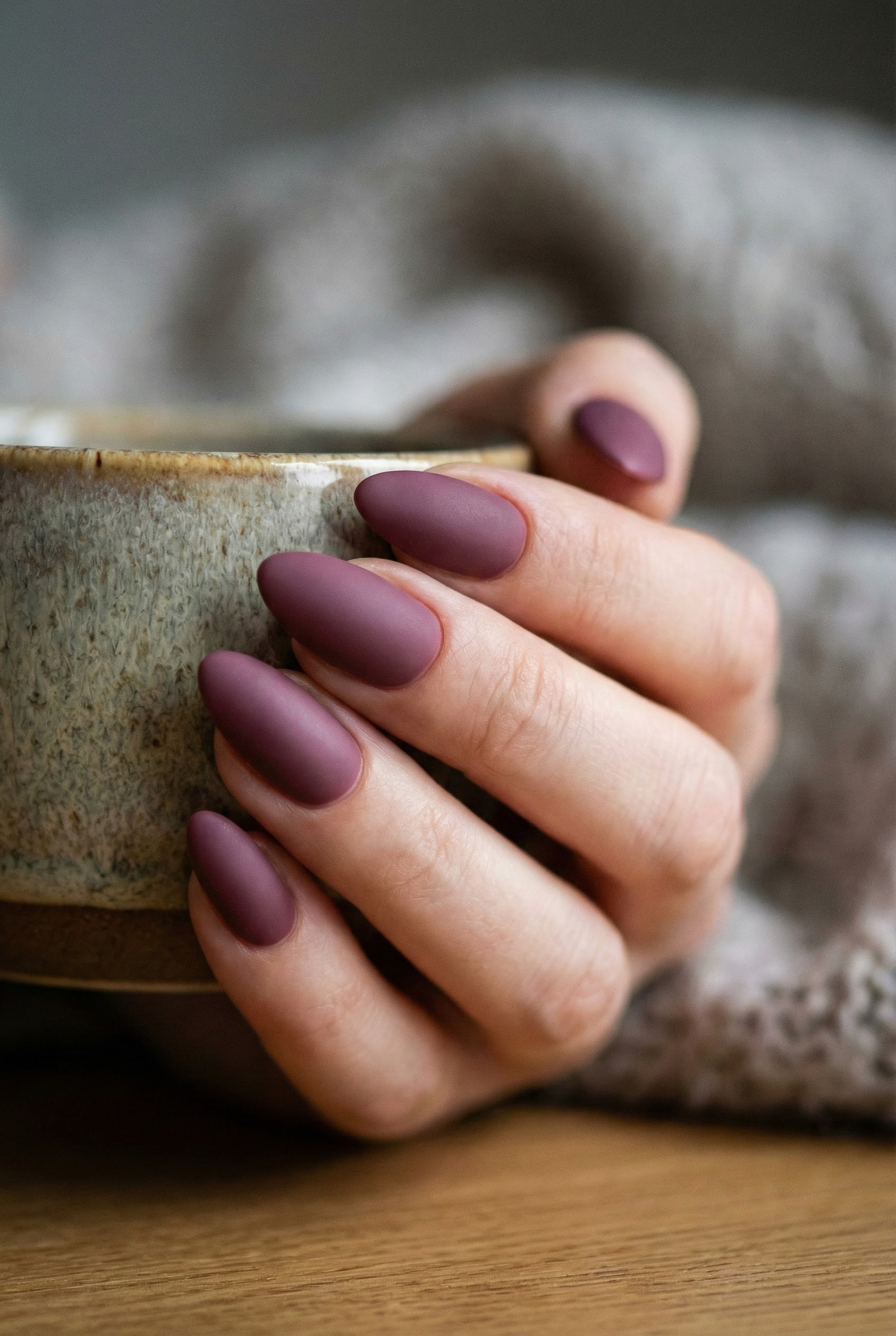 Hand holding a rustic ceramic cup featuring almond-shaped nails in a deep mauve color with a smooth matte finish.