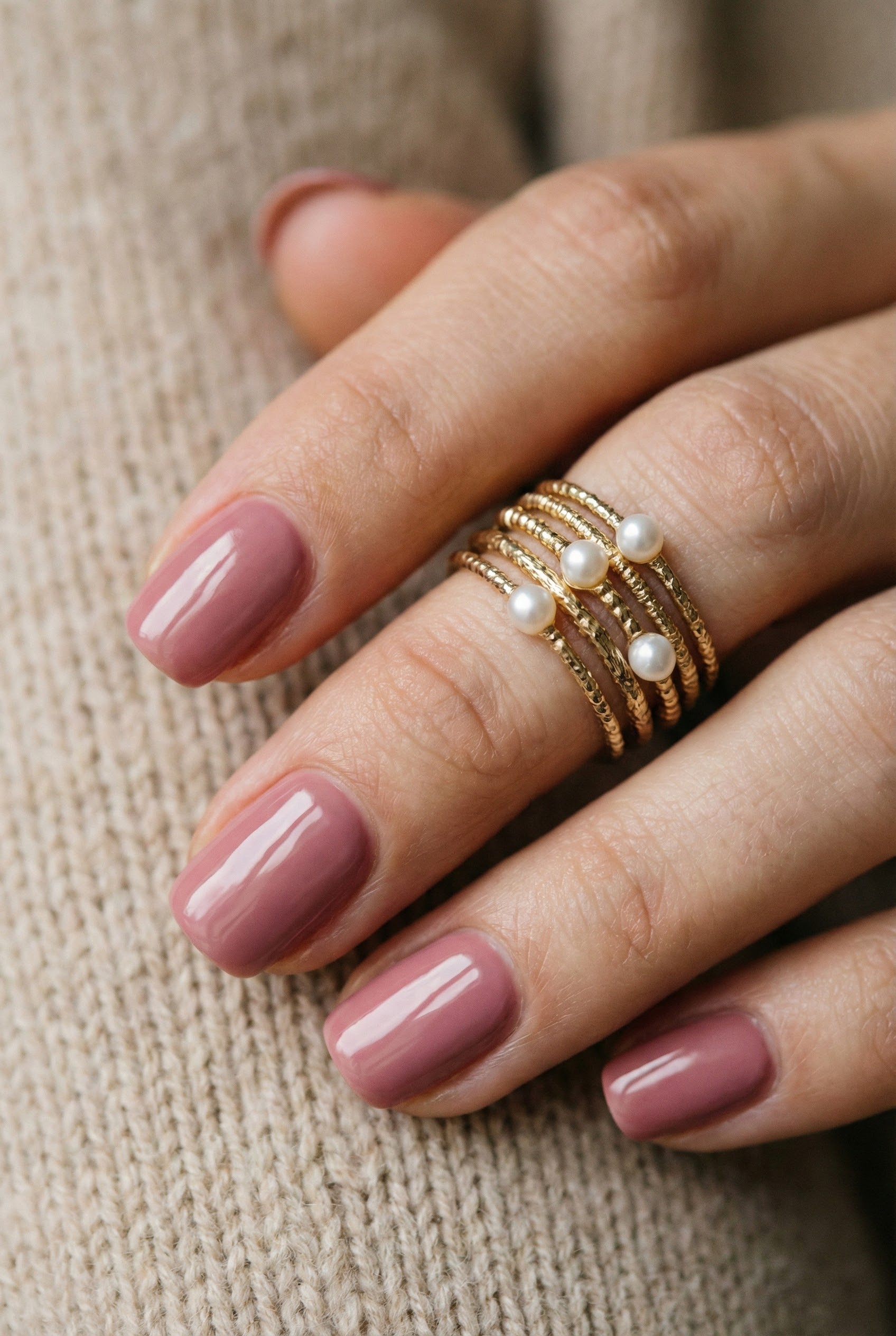 Vertical macro shot of glossy dusty rose nails styled with a stack of gold and pearl rings against a cozy beige knit sweater background.