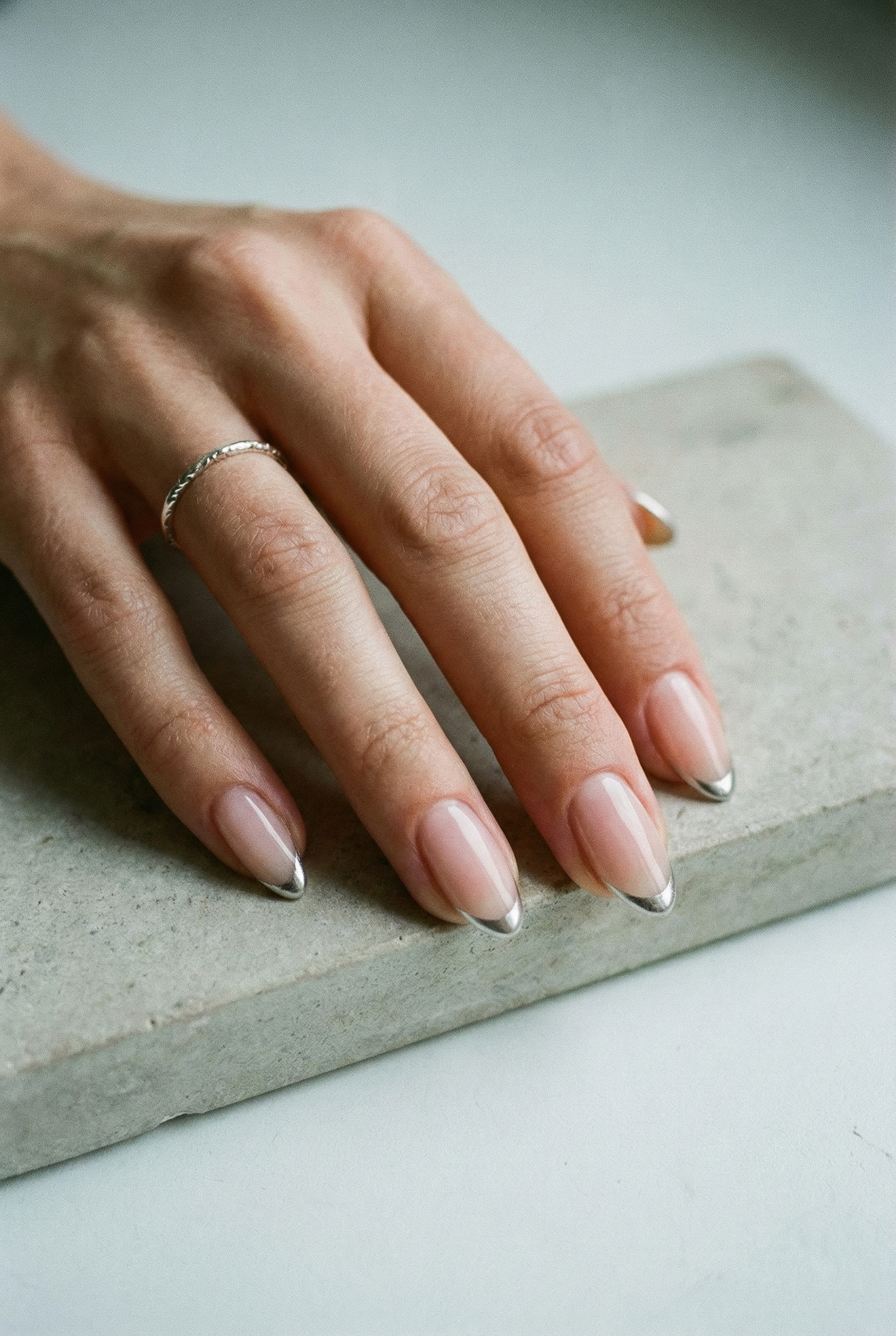 Hand resting on a grey stone surface showcasing a minimalist micro French manicure with thin metallic silver tips.