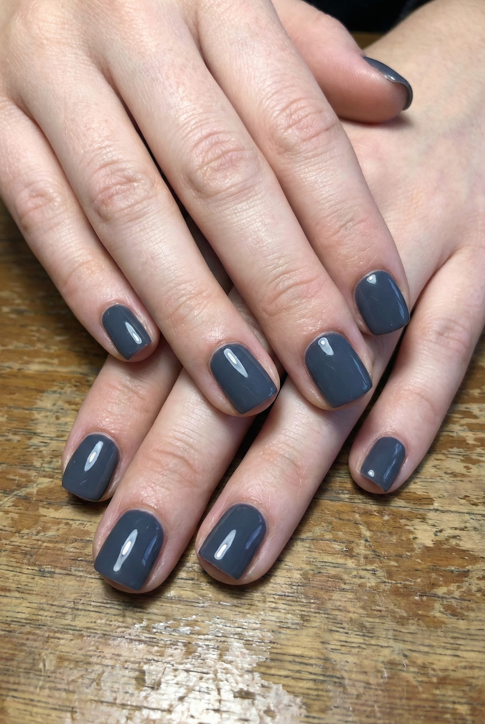 Top-down view of short square nails painted in dark slate grey resting on a wooden desk surface.