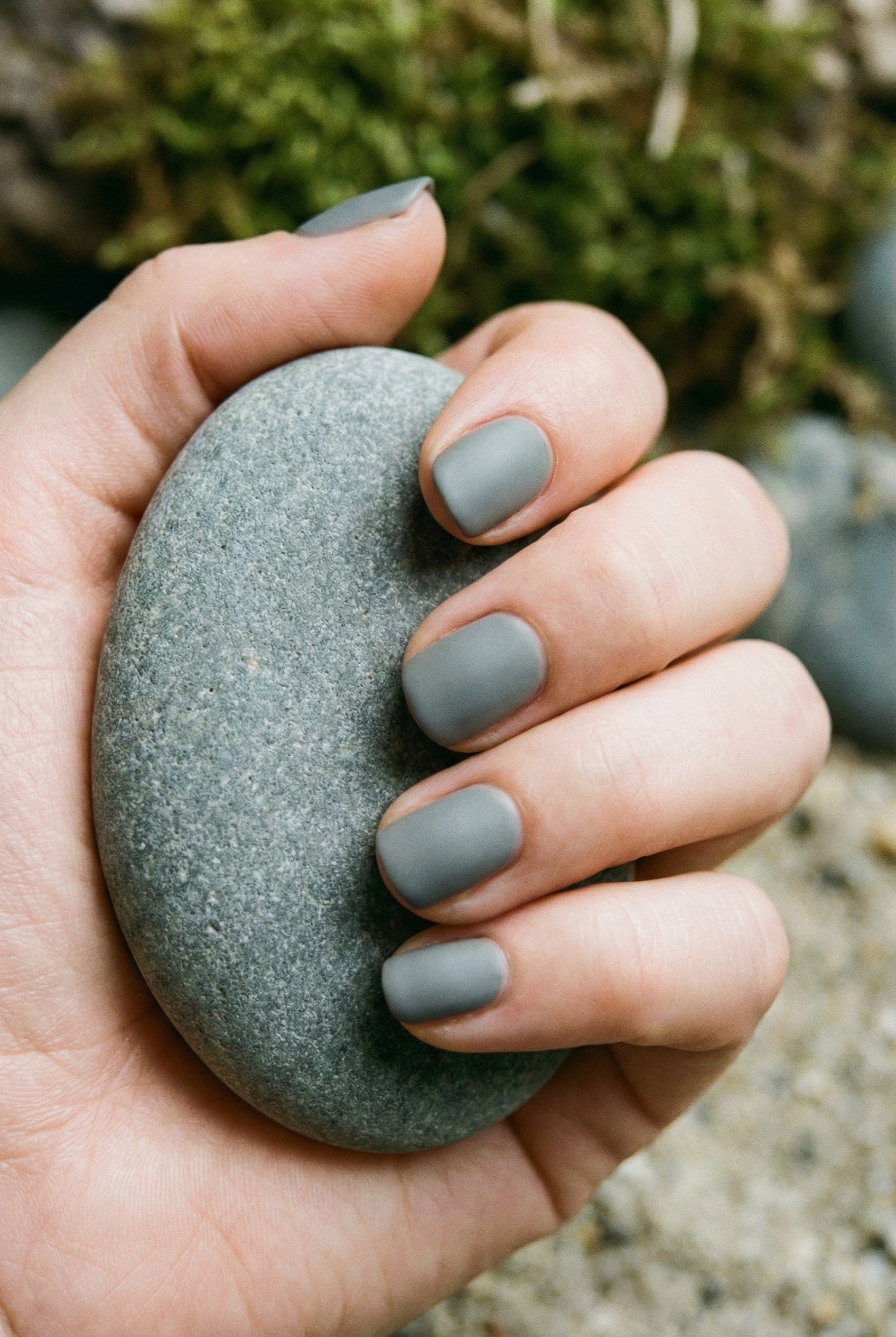 Hand holding a smooth river stone featuring matching matte grey nails that mimic the organic texture.