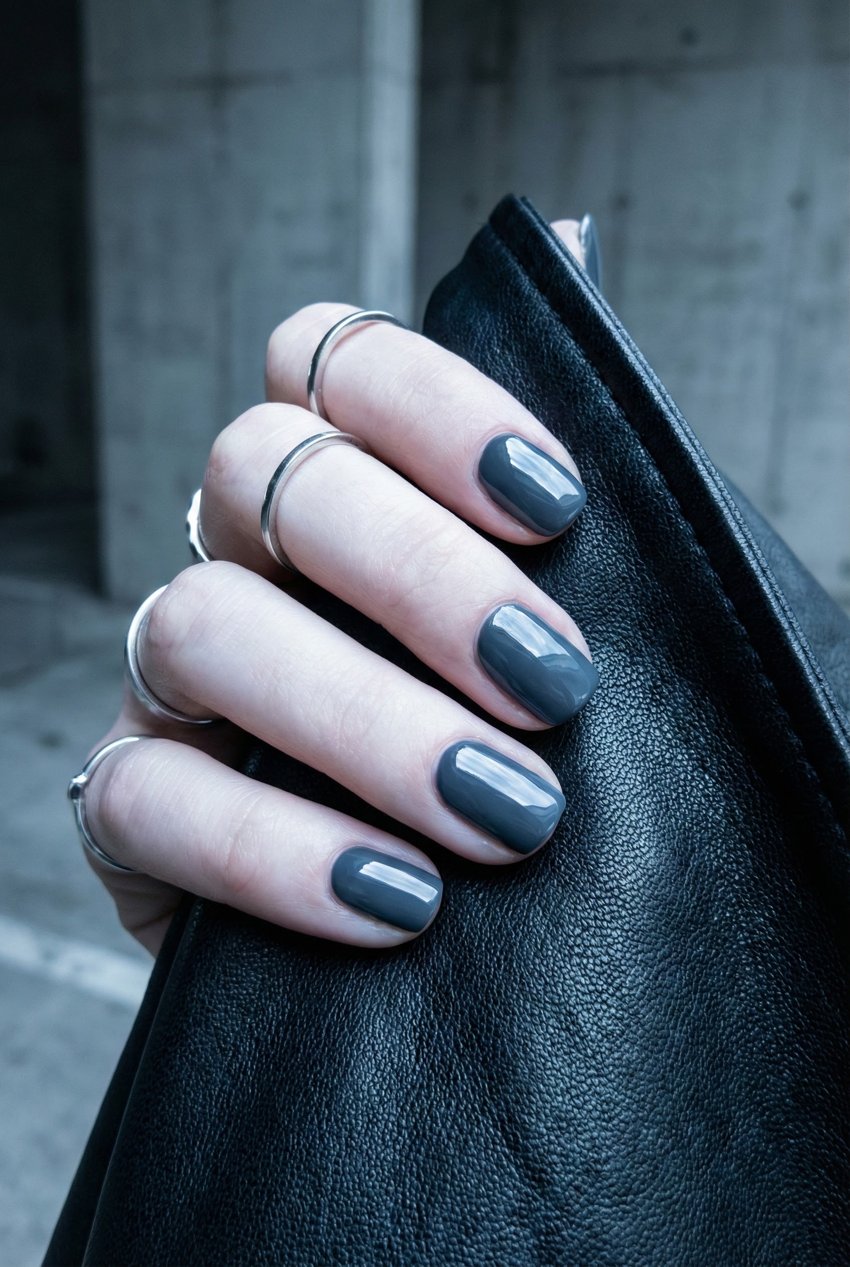 Vertical fashion shot of a hand with glossy slate grey nails holding a black leather bag, accessorized with stacked minimal silver rings.