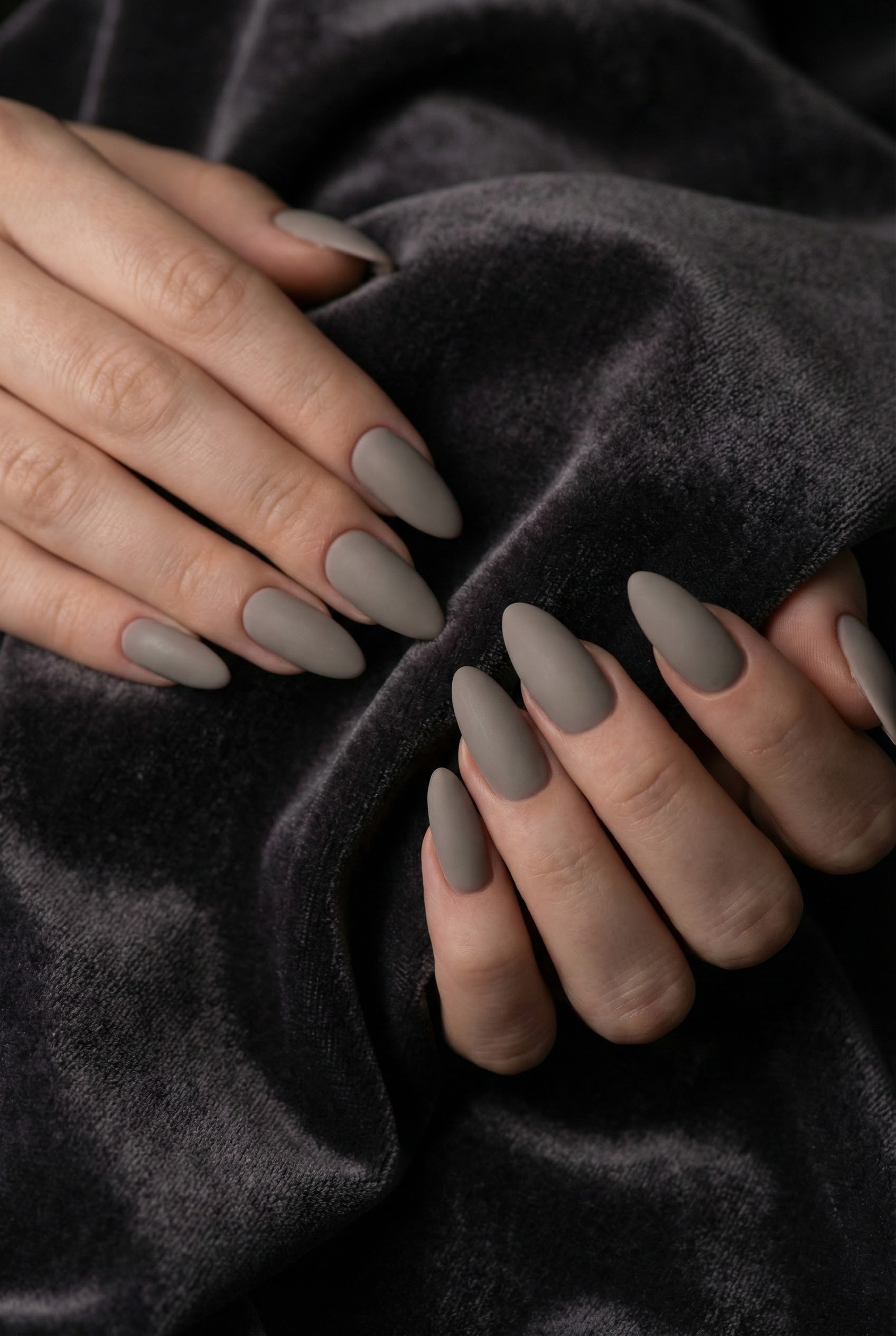 Close-up of matte greige almond-shaped nails resting on dark grey velvet fabric, highlighting the soft, non-shiny texture.