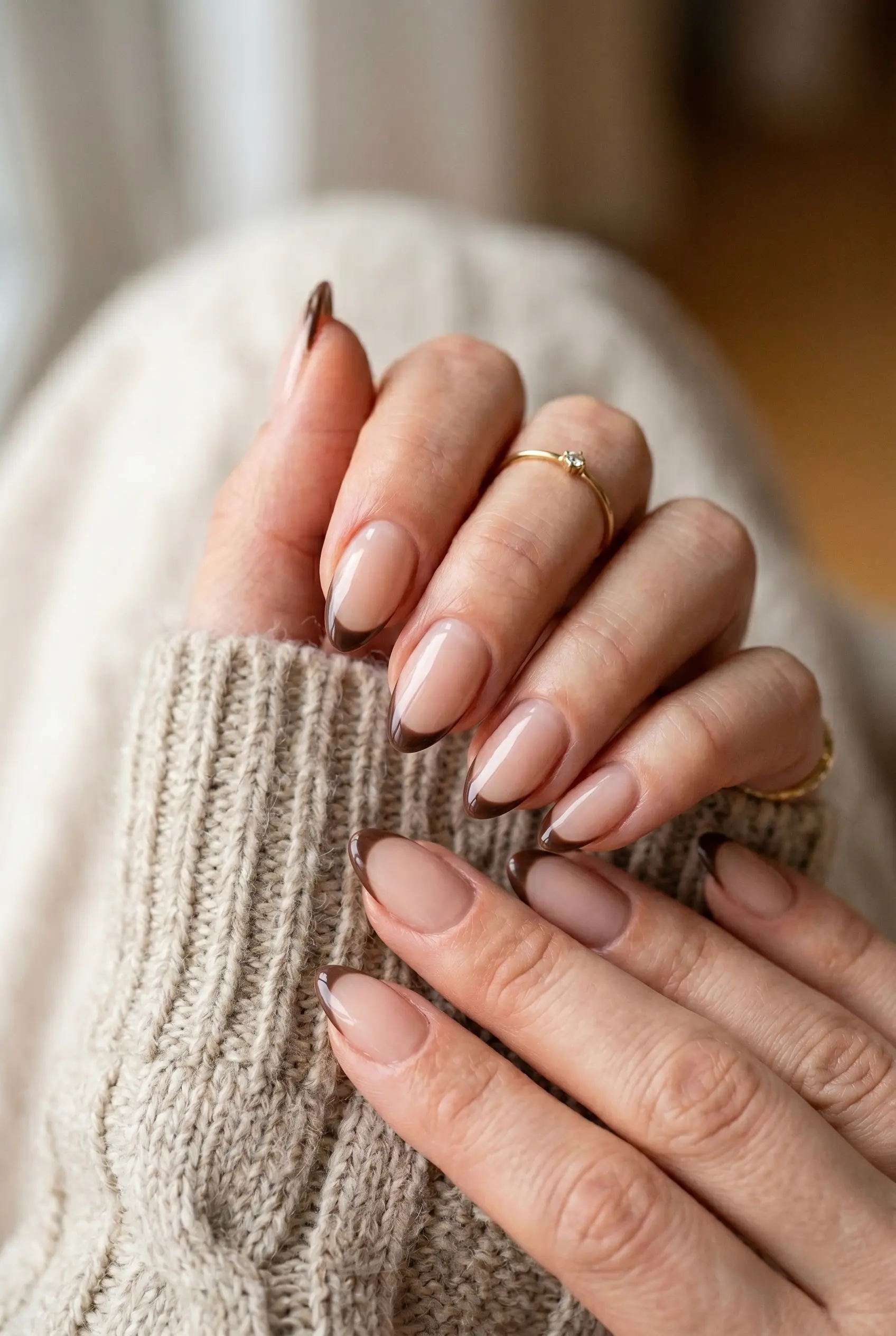An elegant close-up of a brown French manicure with thin mocha tips on a natural nude base, held against a beige knit sweater.