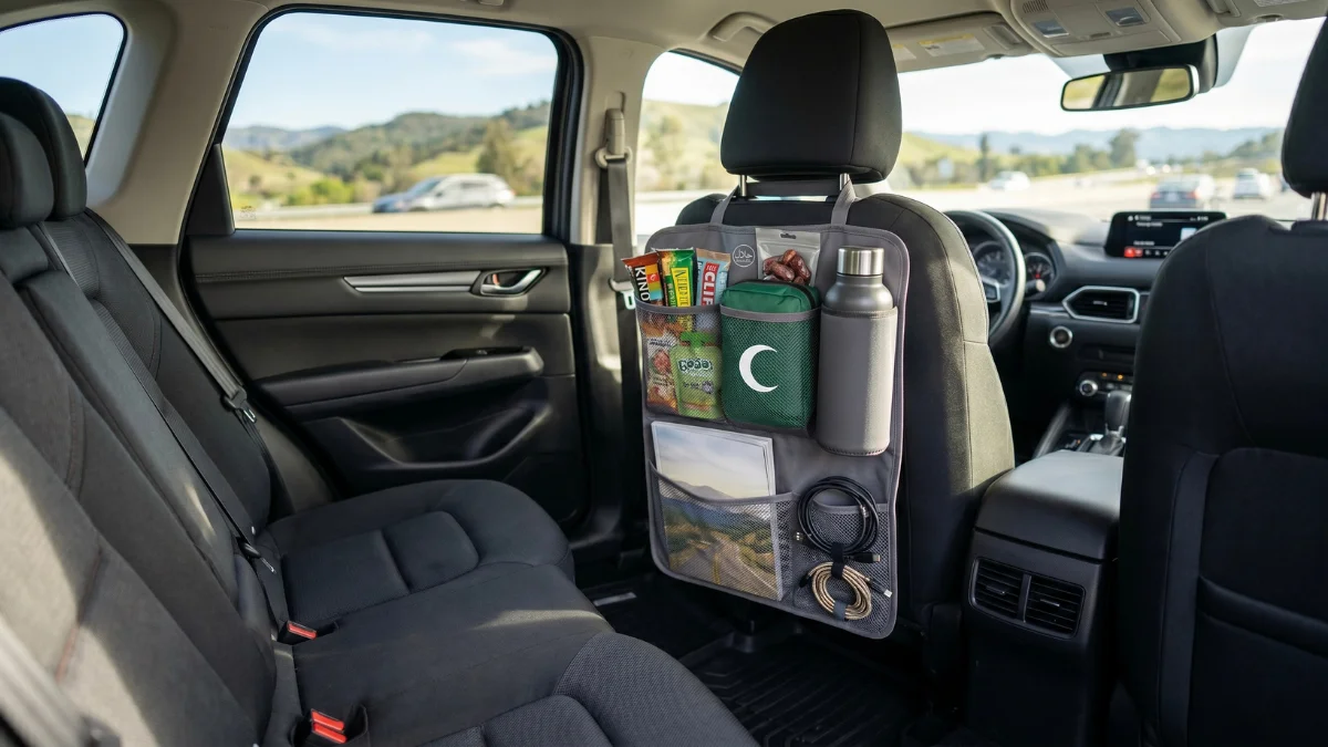A gray multi-pocket organizer hanging on the back of a car seat, neatly holding snacks, a water bottle, a first-aid kit, a magazine, and charging cables for a family road trip.