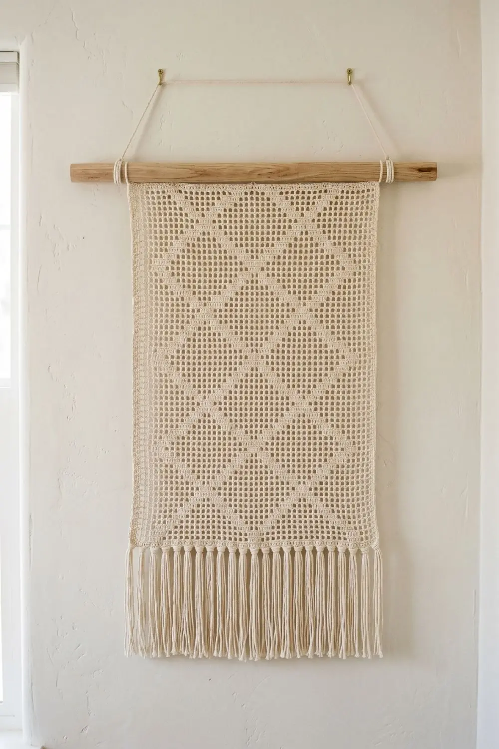 5 Geometric Crochet Wall Hangings That Make a Minimal Home Feel Complete 1 A diamond grid filet crochet wall hanging in undyed natural cotton, suspended from an oak dowel with brass hooks against a white plaster wall, with neat cream fringe at the base