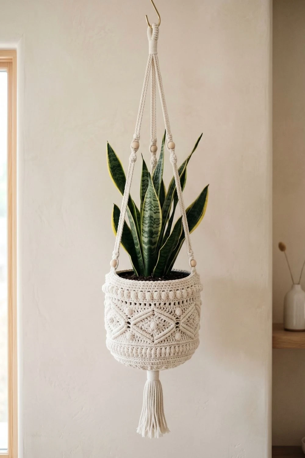 5 Breathtaking Crochet Hanging Baskets to Elevate Your Indoor Garden 2 Elegant cream colored crochet hanging planter with intricate elmas and bobble textures, tassels, and wood beads, holding a vertical snake plant against a pale beige wall.