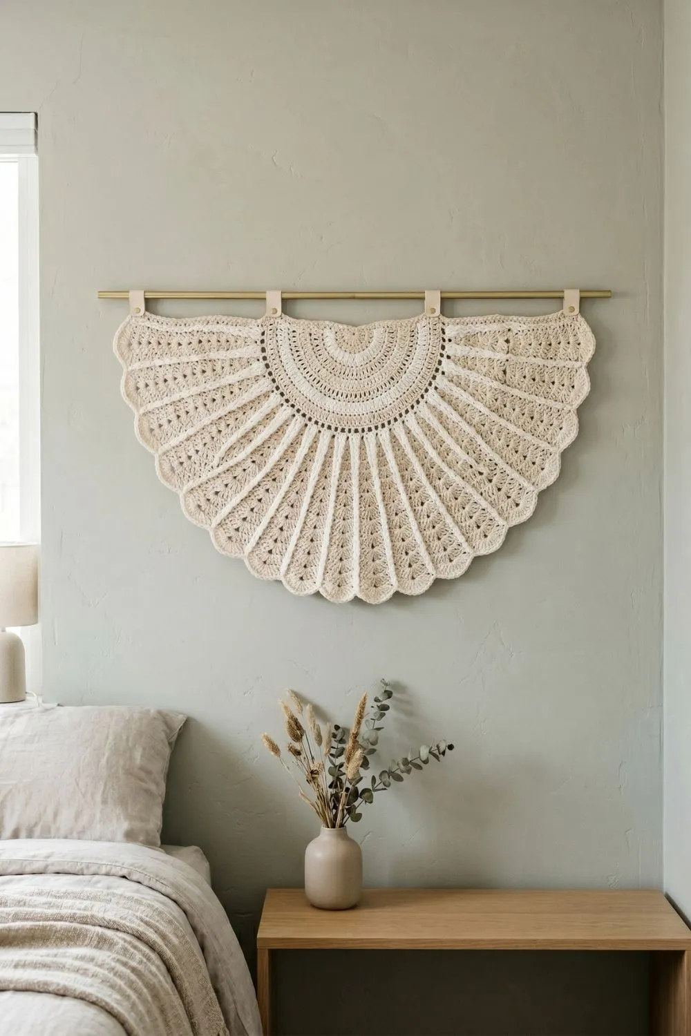 5 Geometric Crochet Wall Hangings That Make a Minimal Home Feel Complete 2 A large half-circle sunburst crochet wall hanging in cream and off-white, mounted on a brass rod against a sage-toned wall in a minimal bedroom, with dried botanicals on a wooden bedside table below