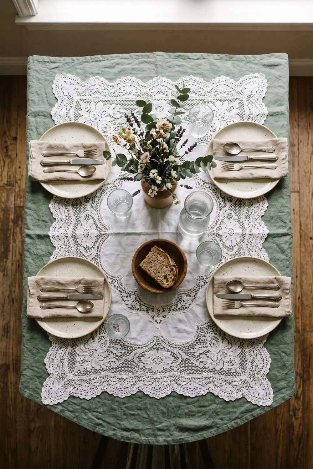 Overhead view of a white lace tablecloth overlay layered on a sage green linen base with rustic place settings