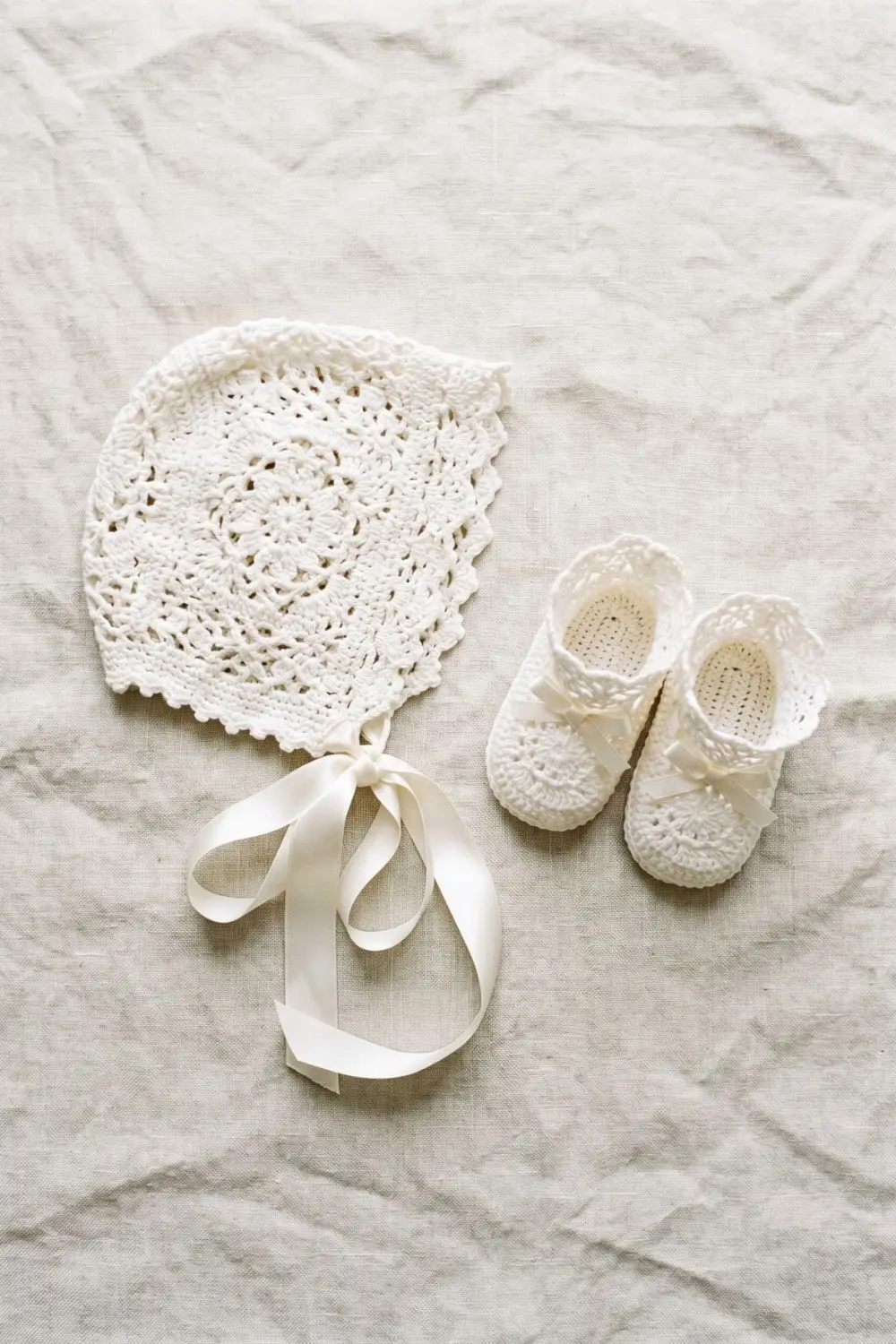 White openwork crochet baby bonnet with long satin tie ribbons beside a pair of matching lace-edge booties on pale linen, soft natural daylight