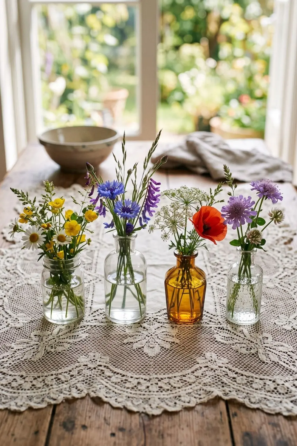 Four mismatched glass and amber jars filled with wildflowers on a crochet lace tablecloth near a window