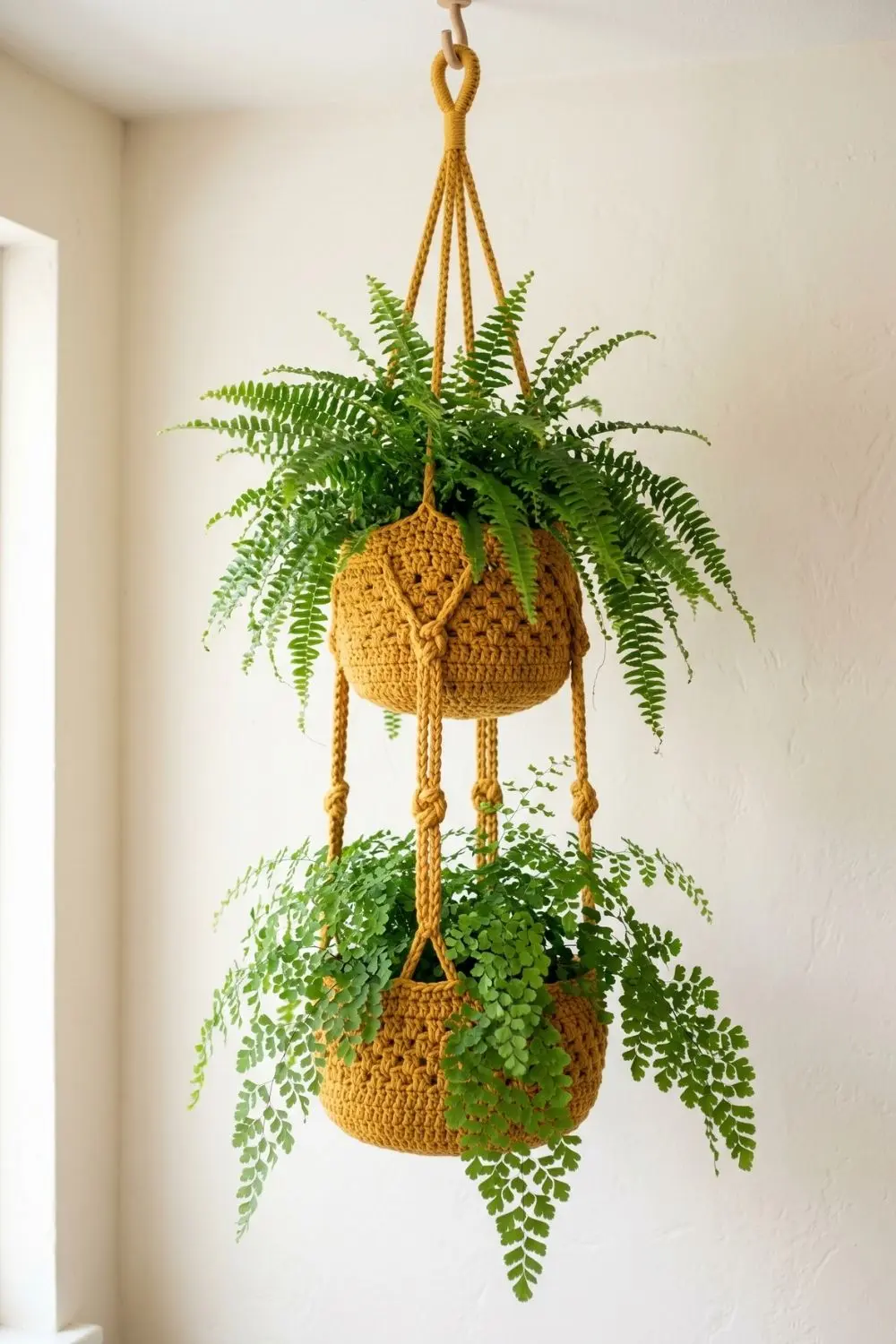 5 Breathtaking Crochet Hanging Baskets to Elevate Your Indoor Garden 3 Two different types of cascading ferns are held within a warm mustard-yellow two-tiered crochet hanging basket structure, suspended by cords against a plain cream wall.