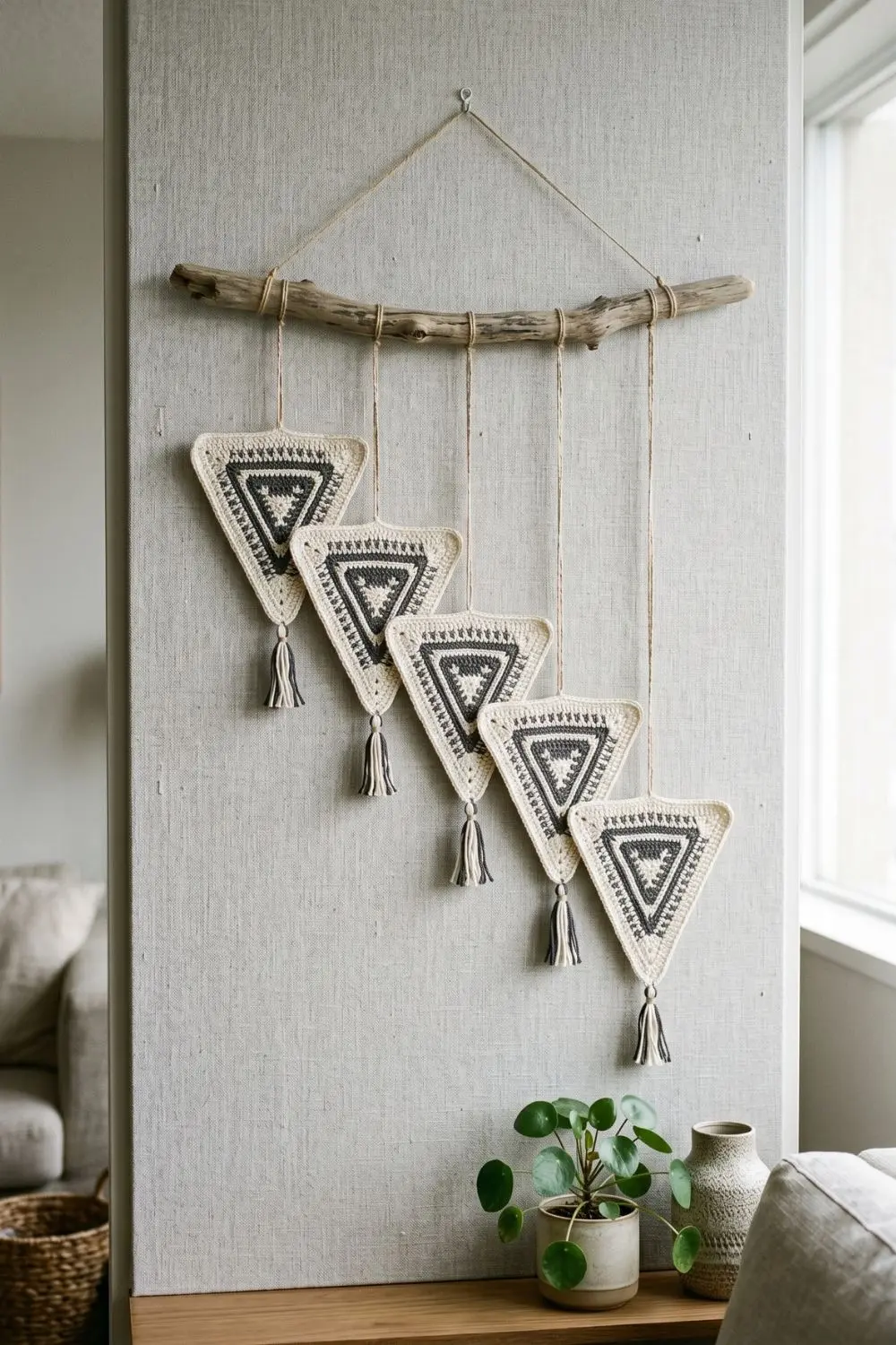 5 Geometric Crochet Wall Hangings That Make a Minimal Home Feel Complete 3 Four ivory and charcoal tapestry crochet triangle motifs with tassels, hanging individually from a natural driftwood branch against a light grey linen wall panel in a minimal living room