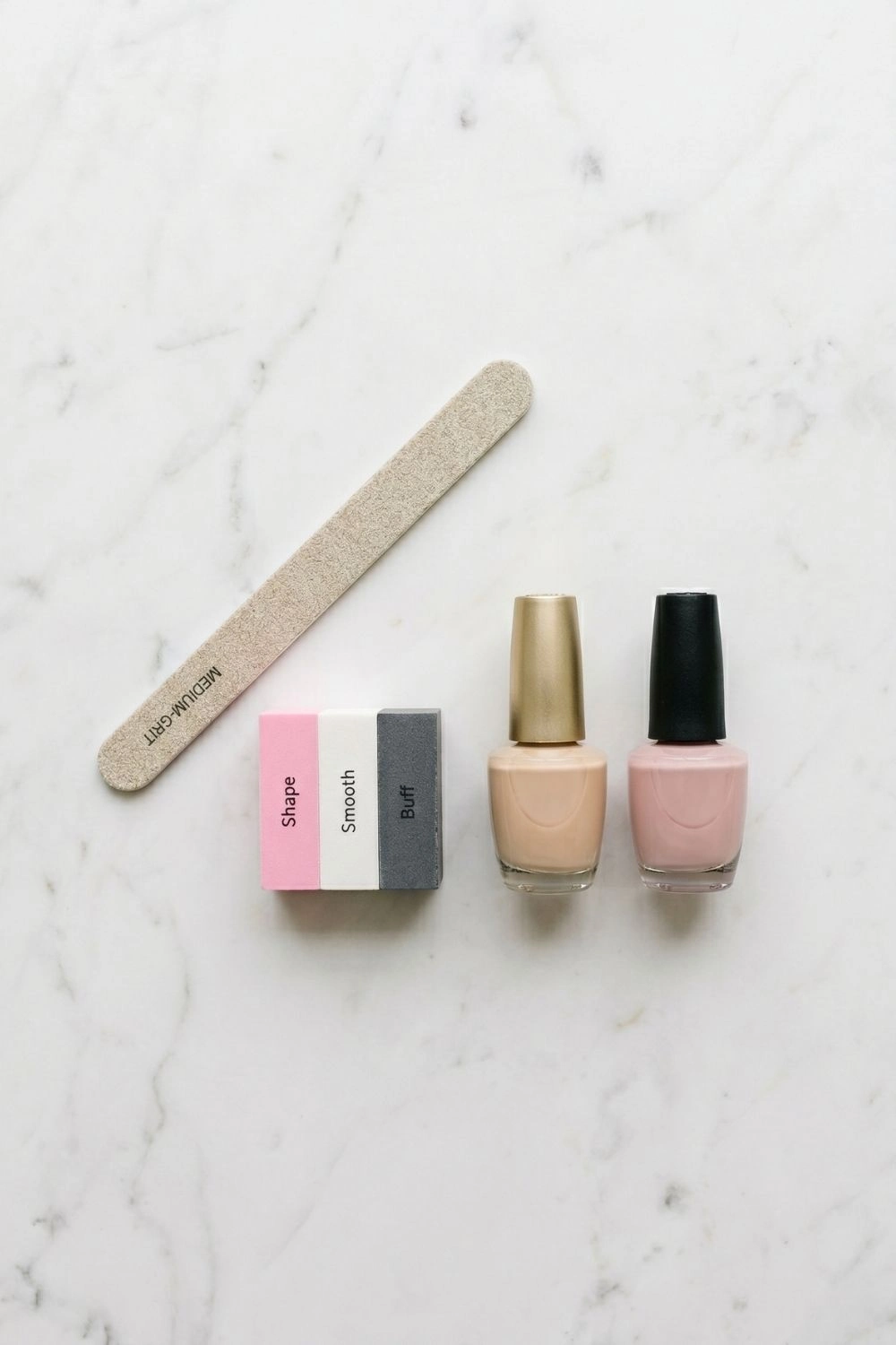 Why Women Are Filing Down Their Long Tips for This Nail Shape 4 Nail file, buffer, and polish bottles arranged on white marble in a flat-lay