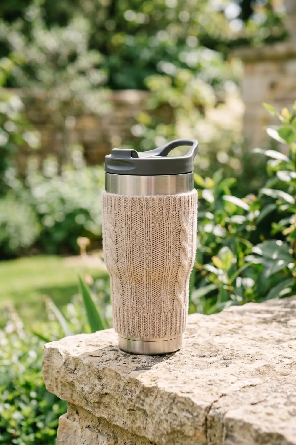 Stainless steel travel tumbler with a beige cable knit sleeve resting on a stone ledge outdoors.