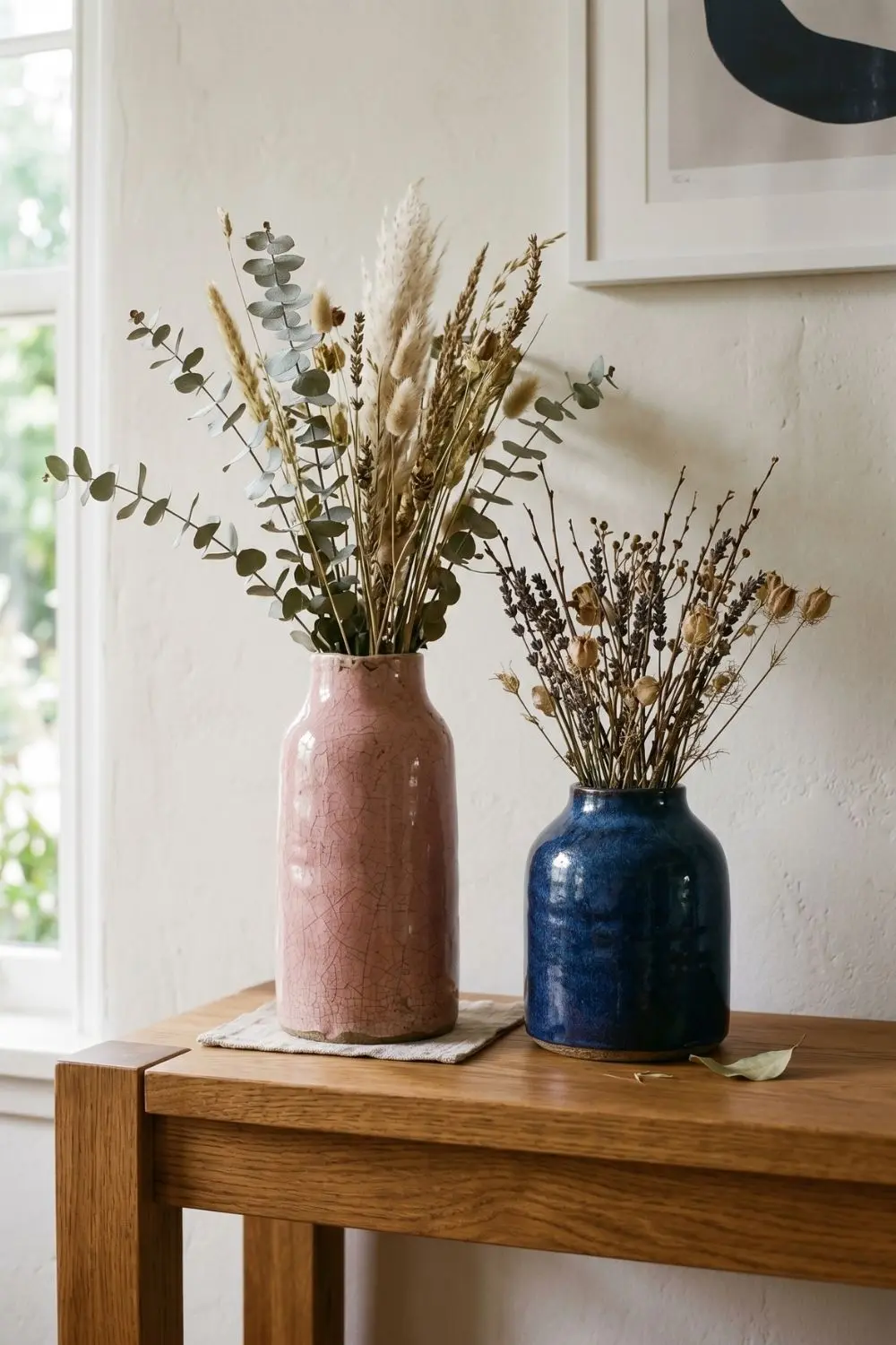 Two ceramic vases on a wood console table, one in a crackled vintage pink glaze and one in a moody blue glaze, both holding dried botanical stems