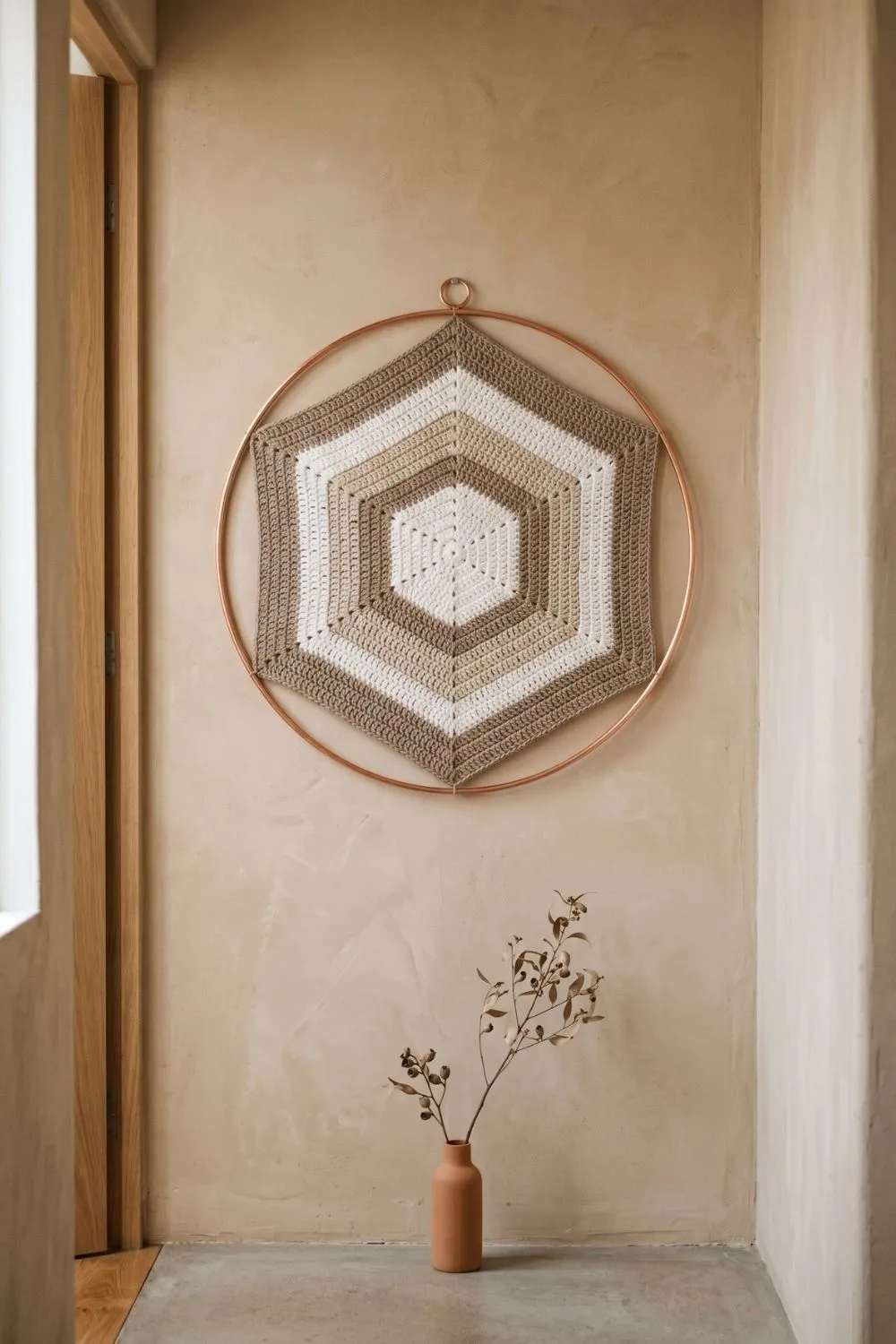5 Geometric Crochet Wall Hangings That Make a Minimal Home Feel Complete 5 A concentric hexagon crochet wall hanging in white, linen, and warm taupe tones, set inside a copper circular frame, hung on a clay-rendered plaster wall in a minimal hallway with a terracotta vase and dried branch below