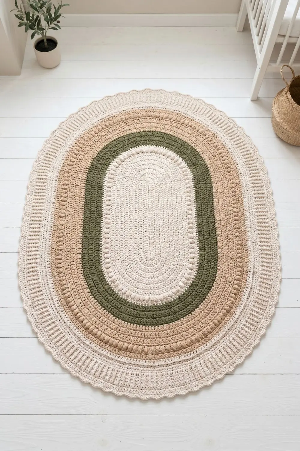 Large oval crochet play mat in warm sand, olive green and undyed natural cotton on a white nursery floor, overhead view