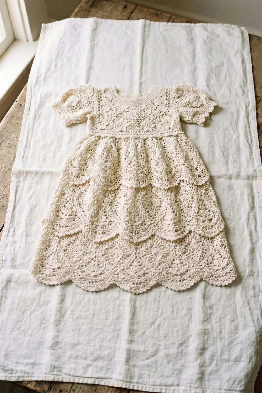 Full-length ivory tiered crochet baby dress with intricate thread lace panels, puff sleeves, and scalloped hem layers, laid flat on white linen on a wooden surface, soft natural window light