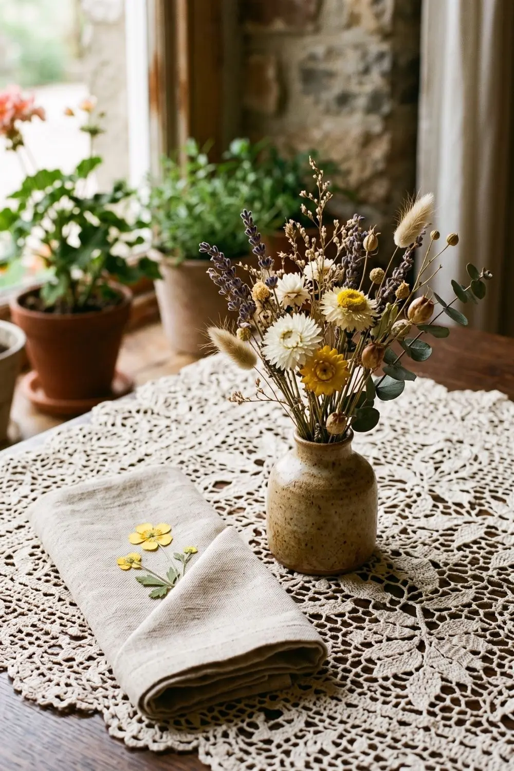 Small ceramic vase of dried lavender and strawflowers on a lace tablecloth beside a folded linen napkin with loose buttercups