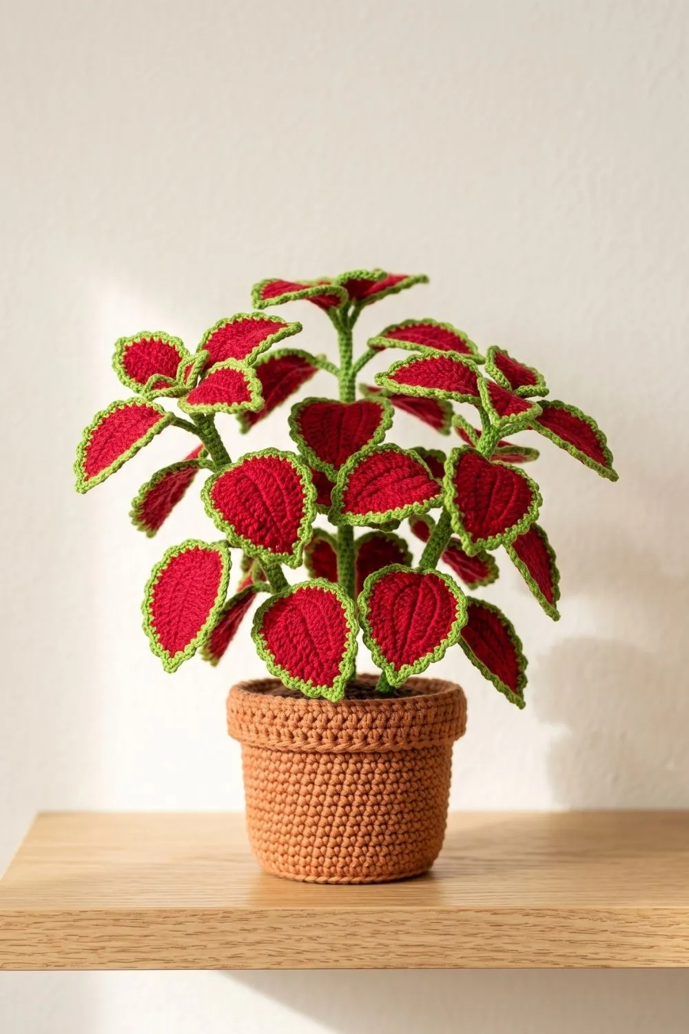 5 Crochet Plant Styles Dominating Pinterest Right Now 4 Full-branched crochet coleus plant in deep crimson red with bright green stems and leaf edging in a terracotta-tone crocheted pot against a white wall