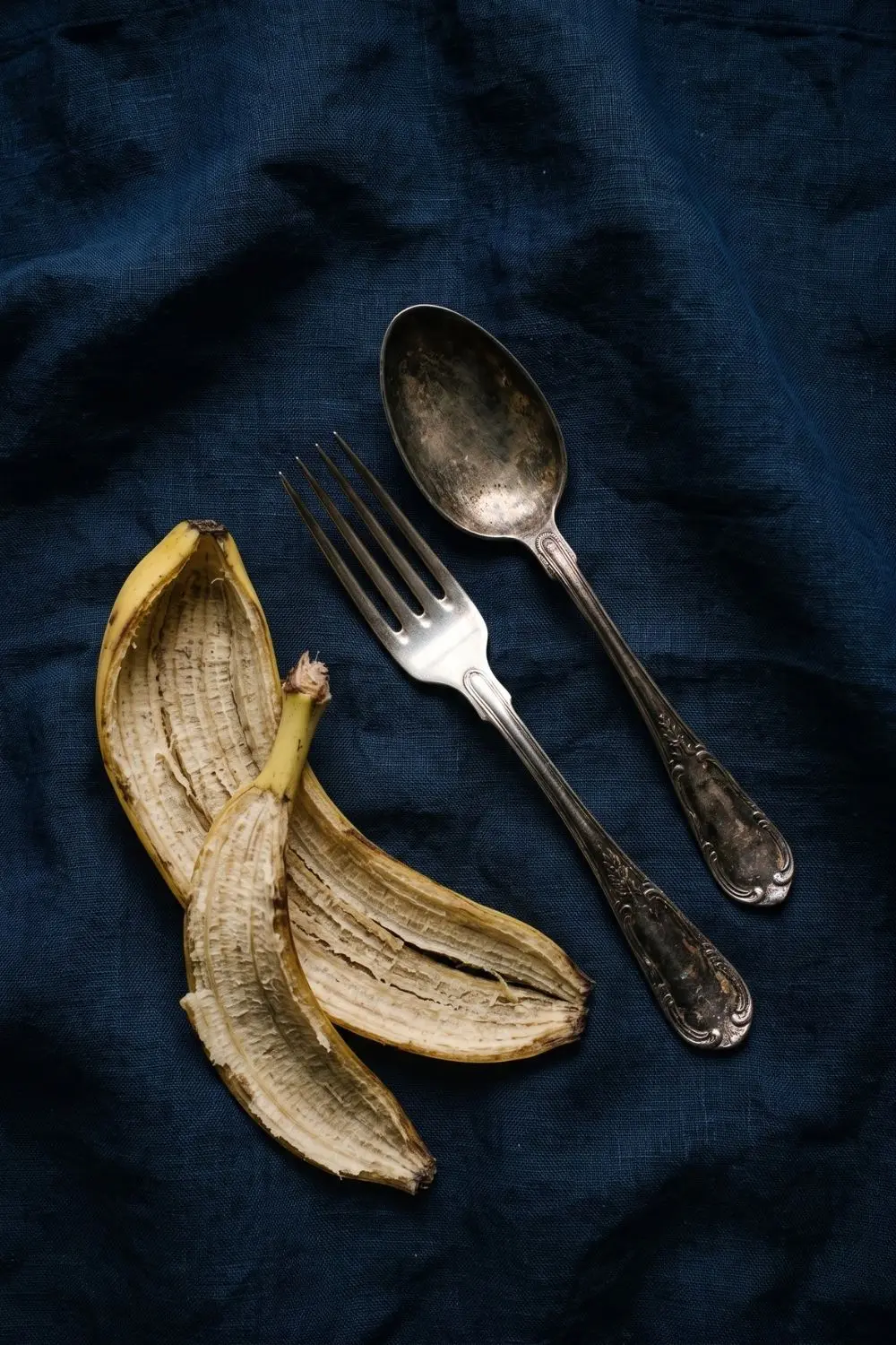What to Do With Banana Peels: Six Practical Uses Around the Home 5 Tarnished silver fork and spoon on a dark cloth beside a fresh banana peel, with one piece showing a polished section